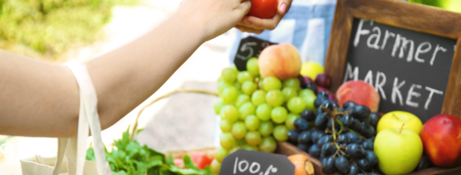 Farmers market produce display—green and black grapes, peaches, apples—with a hand picking up a tomato.